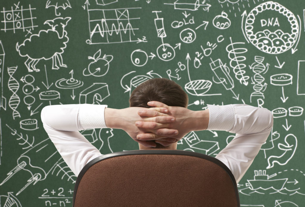 Rear view of man sitting an office chair, with hands behind head, facing a busy blackboard.