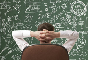 Rear view of man sitting an office chair, with hands behind head, facing a busy blackboard. 