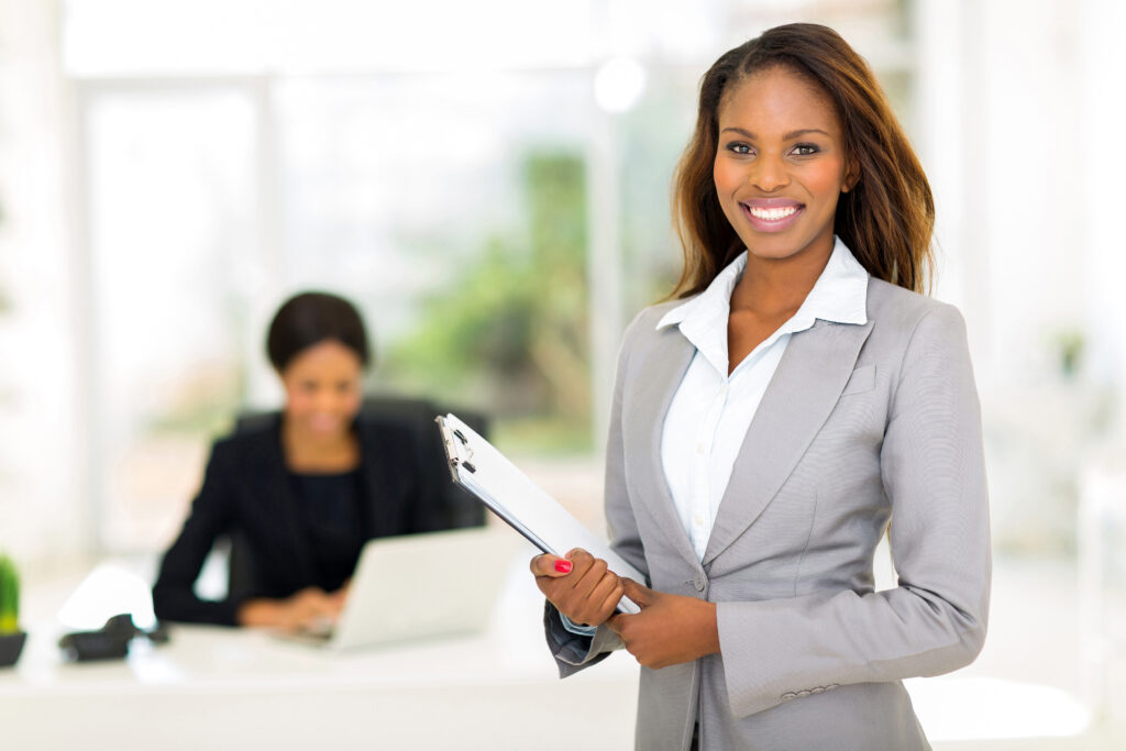 young woman in business suit holding papers, smiling, with another woman in background, working at a desk